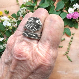 May include: Two rings with a black and white floral pattern are displayed on a hand. The rings have a distinctive, artistic design. The background features flowers and foliage, adding a natural element. The rings are a statement piece.