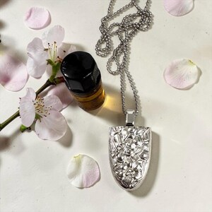 Puede incluir: Un collar con colgante plateado con un diseño floral, sobre una superficie blanca. Una pequeña botella de vidrio con tapa negra y líquido ámbar está al lado. También hay pétalos de flores rosas y una rama con flores blancas y rosas.