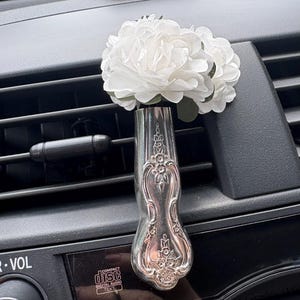 Puede incluir: Un clip de ventilación para coche de metal plateado con forma de mango de cuchara vintage, con intrincados patrones florales en relieve. La parte superior del clip sostiene un grupo de flores artificiales blancas con hojas verdes.