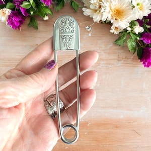 May include: A large silver safety pin with an ornate floral design on the head.