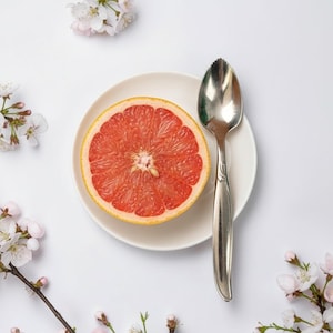 Könnte beinhalten: Eine halbierte rosa Grapefruit liegt auf einem kleinen weißen Keramikteller neben einem silbernen gezackten Grapefruitlöffel. Der Teller ist von zarten weißen Kirschblütenzweigen mit kleinen rosa Knospen umgeben, alles auf einer weißen Fläche.