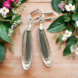 May include: A pair of silver dangle earrings made from repurposed silverware. The earrings feature a decorative design and are suspended from silver hooks.