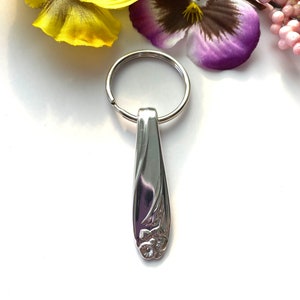 Könnte beinhalten: Ein silberner Schlüsselanhänger mit einem Blumendesign. Der Schlüsselanhänger ist aus einem wiederverwendeten Besteckteil gefertigt.