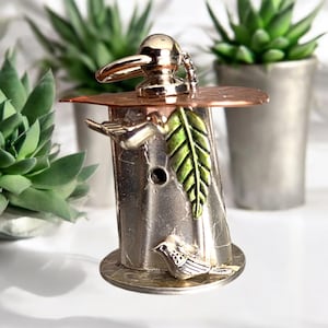 Puede incluir: Un amuleto de plata en forma de casita de pájaros con un techo de cobre, una hoja verde y un pequeño pájaro de plata. El amuleto tiene un anillo de plata para sujetarlo a un llavero.