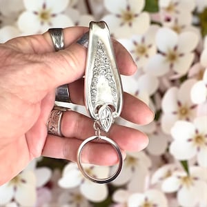 May include: Silver spoon keychain with ornate floral detailing. The spoon's handle is repurposed into a keychain, featuring a decorative design and a silver key ring. The background is blurred with white flowers.
