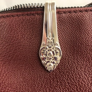 May include: A burgundy leather pouch with a silver spoon handle as a zipper pull. The spoon handle has floral and scrollwork detailing. The zipper is black.