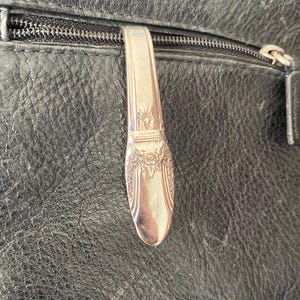 May include: A black leather bag with a silver zipper and a silver metal pull tab. The pull tab is shaped like a spoon and has a decorative design.
