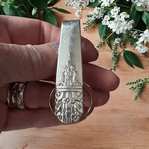 Könnte beinhalten: Silberfarbener Schlüsselanhänger aus einer wiederverwerteten Gabel. Der Gabelgriff hat ein verziertes Blumendesign. Ein runder Schlüsselring ist am unteren Ende des Gabelgriffs befestigt. Der Schlüsselanhänger wird vor einem Holzuntergrund mit weißen Blumen im Hintergrund gehalten.