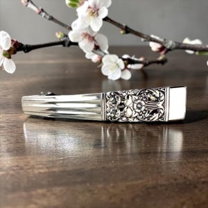 Puede incluir: Un pitillera plateada rectangular con detalles florales ornamentados. La pitillera tiene un diseño acanalado en el cuerpo y un extremo rectangular blanco en blanco. Una ramita de flores blancas está en el fondo.