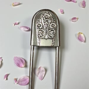 May include: A silver-toned safety pin with an ornate floral design on the head. The pin is surrounded by scattered pink flower petals on a white background. The pin's design features detailed flowers and scrollwork.