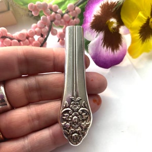 May include: A silver-toned metal handle featuring an embossed floral pattern at the base. The handle has a smooth, tapered upper section and a rounded, decorative lower end with intricate rose and leaf detailing in a polished metallic finish.