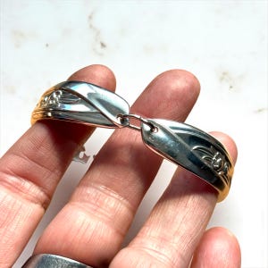 Puede incluir: Un brazalete plateado con un diseño floral, elaborado con cubiertos reciclados. El brazalete presenta un diseño articulado con un cierre de conexión. El metal tiene un acabado pulido, que refleja la luz. El brazalete es sostenido por una mano.