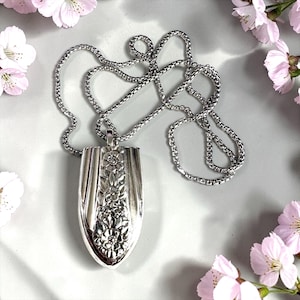 May include: A silver pendant necklace featuring a floral design. The pendant is shield-shaped with embossed flowers and leaves. The chain is a delicate, textured link. Pink flowers are in the background.