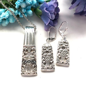 Pode incluir: Um conjunto de colar e brincos de prata com um design floral. O colar tem uma corrente e um pingente com um padrão floral. Os brincos são brincos pendurados com um design floral.