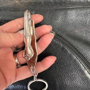 Könnte beinhalten: Besteck-Schlüsselanhänger aus einem Löffel mit floralem Design. Der Löffel ist an einem silbernen Schlüsselring befestigt. Der Hintergrund ist eine schwarze Lederoberfläche.