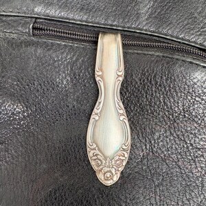 May include: A silver spoon used as a zipper pull on a black leather bag. The spoon has an ornate floral design at the base and decorative edges. The zipper is black.