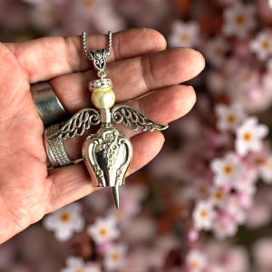 May include: Silver-toned angel pendant necklace. The pendant features a pearl, iridescent accents, and detailed wings. The pendant's body has a floral design. The necklace is held in a hand.