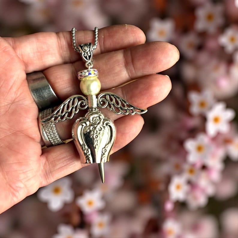 May include: Silver-toned angel pendant necklace. The pendant features a pearl, iridescent accents, and detailed wings. The pendant's body has a floral design. The necklace is held in a hand.