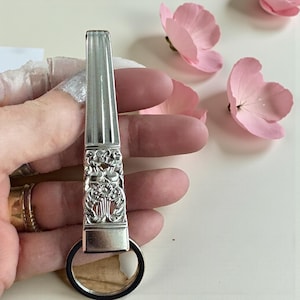 Puede incluir: Un llavero plateado con un diseño floral decorativo. El llavero tiene una parte superior rectangular con líneas verticales y un anillo circular en la parte inferior. Flores rosas en el fondo.