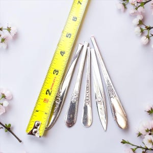May include: Five silver-toned metal spoon handles with various embossed floral patterns arranged vertically next to a yellow Stanley measuring tape showing a length of approximately 7 inches for the longest handle.