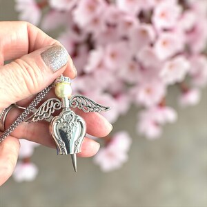 May include: A silver-toned angel pendant necklace. The pendant features a pearl head, a crown, and detailed wings. The angel's body has an ornate design. The necklace is held against a blurred background of pink flowers.