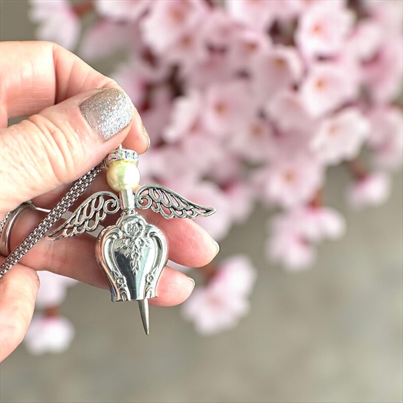 May include: A silver-toned angel pendant necklace. The pendant features a pearl head, a crown, and detailed wings. The angel's body has an ornate design. The necklace is held against a blurred background of pink flowers.