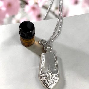 May include: A silver pendant necklace with a rectangular, floral-detailed pendant. The pendant is attached to a delicate chain. A small, black-topped bottle is next to the necklace. The background is blurred with pink flowers.