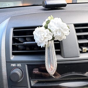 May include: A silver-toned metal car air vent clip shaped like a small vase, featuring an ornate handle design and filled with three artificial white flowers with green leaves, attached to the horizontal slats of a vehicle dashboard air vent.
