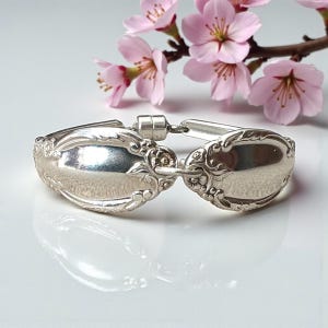 Puede incluir: Un brazalete plateado hecho de cubiertos reciclados. El brazalete presenta detalles ornamentados y está conectado por un cierre. Las flores de cerezo rosas en el fondo añaden un toque de color y elegancia a la joya.