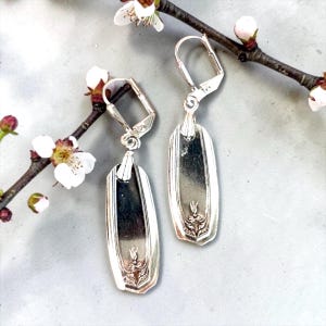 May include: A pair of silver-toned dangle earrings featuring elongated, rectangular metal components with rounded corners and a decorative embossed floral motif at the base. The earrings are attached to silver-toned lever-back ear wires.