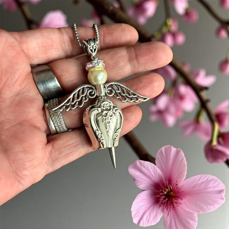 May include: A silver-toned angel pendant necklace with a pearl and rhinestone accents. The pendant features detailed wings and a decorative body. The necklace is displayed against a backdrop of pink flowers.