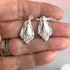 Può includere: Un paio di orecchini pendenti color argento con un design decorativo e ornato che ricorda i manici delle posate vintage. Ogni orecchino presenta una finitura lucida e riflettente con dettagli floreali in rilievo alla base e un piccolo occhiello.