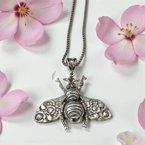 May include: A silver-toned bee pendant necklace. The bee pendant has intricate floral detailing on its wings and body. The necklace is displayed on a white surface, surrounded by pink flower petals.