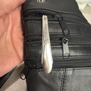 Puede incluir: Bolso bandolera de cuero negro con múltiples compartimentos con cremallera. Una cuchara plateada con un diseño floral está sujeta a la cremallera superior. El bolso tiene una correa y un anillo metálico.