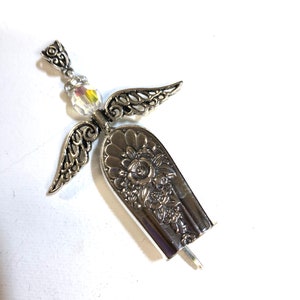 May include: A handcrafted silver angel pendant. The angel features detailed wings, a floral design on the body, and a shimmering crystal bead. The pendant is made from repurposed silverware, showcasing a unique, vintage aesthetic.
