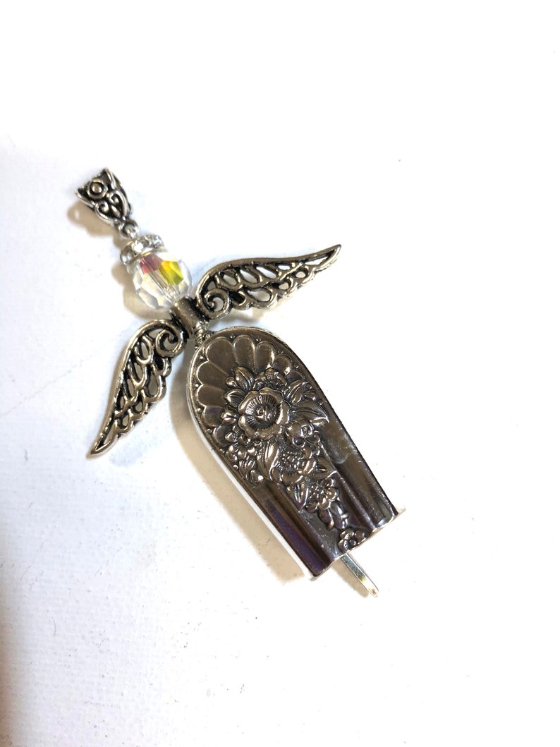 May include: A handcrafted silver angel pendant. The angel features detailed wings, a floral design on the body, and a shimmering crystal bead. The pendant is made from repurposed silverware, showcasing a unique, vintage aesthetic.