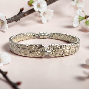 Puede incluir: Un brazalete plateado con superficie texturizada y detalles florales. El brazalete tiene un cierre magnético y se muestra sobre un fondo rosa con flores y ramas blancas.