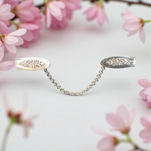May include: Silver collar clip with an ornate floral design on each end, linked by a delicate chain. The clip is set against a soft white background, with pink cherry blossoms in the foreground and background.