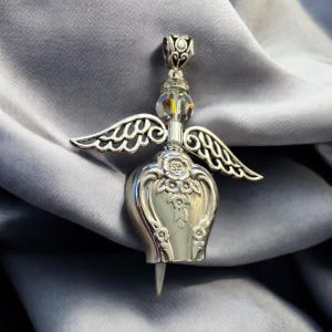 May include: Silver angel pendant featuring a heart-shaped charm with wings. The charm is decorated with floral designs and a crystal bead hangs from the top.