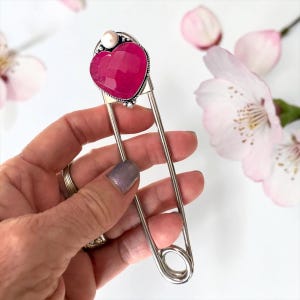 May include: A large silver-toned safety pin brooch featuring a faceted, heart-shaped hot pink gemstone at the top. A small, round, white pearl-like bead is set directly above the heart, framed by decorative silver metalwork detailing.