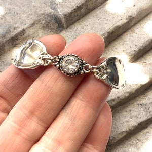 May include: A silver bracelet made from two spoon handles connected by a silver chain with a clear rhinestone in the center.
