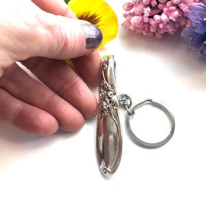 Puede incluir: Un llavero de cuchara de plata con un diseño floral y un anillo de llave de plata.