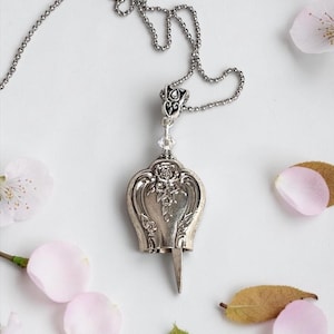 May include: A silver-toned bell pendant necklace featuring an ornate floral embossed design on the surface. The bell is suspended from a delicate ball chain and includes a small clear crystal accent above the bell, which has a pointed clapper.