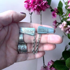 May include: A pair of silver spoon-shaped jewellery pieces connected by a silver chain. The pieces are decorated with floral designs.
