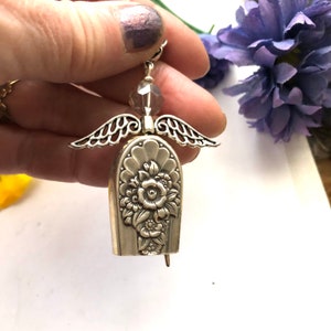 May include: Silver-toned angel earring with intricate wing details and a floral design on the body. The angel's head features a faceted bead. The earring is held up by a silver hook.