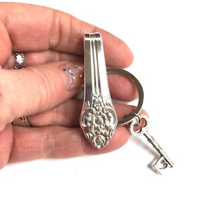 Puede incluir: Un llavero plateado con un dije en forma de cuchara con detalles florales y un dije en forma de llave. El dije de cuchara está unido a un anillo de metal, y el dije de llave tiene un recorte en forma de corazón. Un accesorio único.