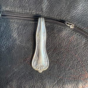 Könnte beinhalten: Ein silberner Löffel als Reißverschlussanhänger an einer schwarzen Ledertasche. Der Löffel ist mit einem dekorativen floralen Design verziert. Der Reißverschluss ist schwarz und die Tasche hat eine strukturierte Oberfläche.