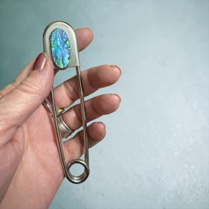 Peut inclure: Une grande épingle de sûreté argentée avec une incrustation ovale en coquille d'ormeau. La coquille présente des reflets bleus, verts et violets. L'épingle a une extrémité enroulée et un fermoir sûr, adapté à divers usages.