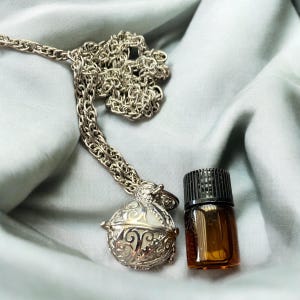 Op de afbeelding: Een zilveren ketting met een decoratieve zilveren bolvormige hanger en een klein bruin glazen flesje met een zwarte dop.