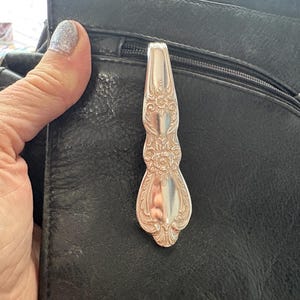Puede incluir: Un mango de cuchara de metal plateado con un intrincado patrón floral y de volutas en relieve. El mango está curvado en la parte superior y se ensancha hacia la base, mostrando una superficie pulida y reflectante con grabados decorativos detallados.